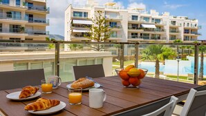 Une table de petit-déjeuner avec des croissants, du jus d’orange et un panier de fruits, placée sur un balcon donnant sur une piscine et des bâtiments modernes.