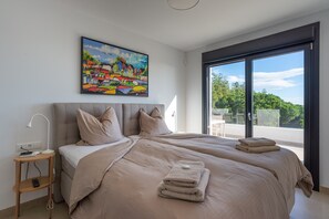 A bedroom with a large bed, a bedside table with a lamp, a painting on the wall, and a view through the window.