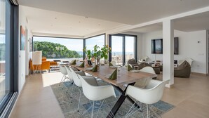 A spacious dining area with a long table, white chairs, and a view of greenery through large windows. - Luxury Villa with Private Pool, with Sea View, Wi-Fi and Air Conditioning (La Cala de Mijas)