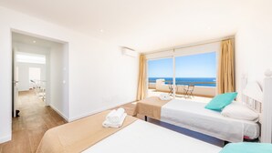 Habitación de hotel con dos camas, un balcón con vista al mar y una sala de estar separada con una mesa y sillas.