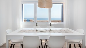 Un comedor con una mesa blanca, sillas y vistas al mar a través de amplios ventanales.