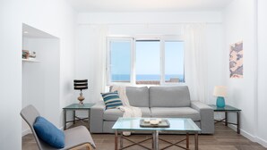 Un salón moderno con un sofá, una mesa de centro de vidrio, una silla y vistas al mar a través de amplios ventanales.