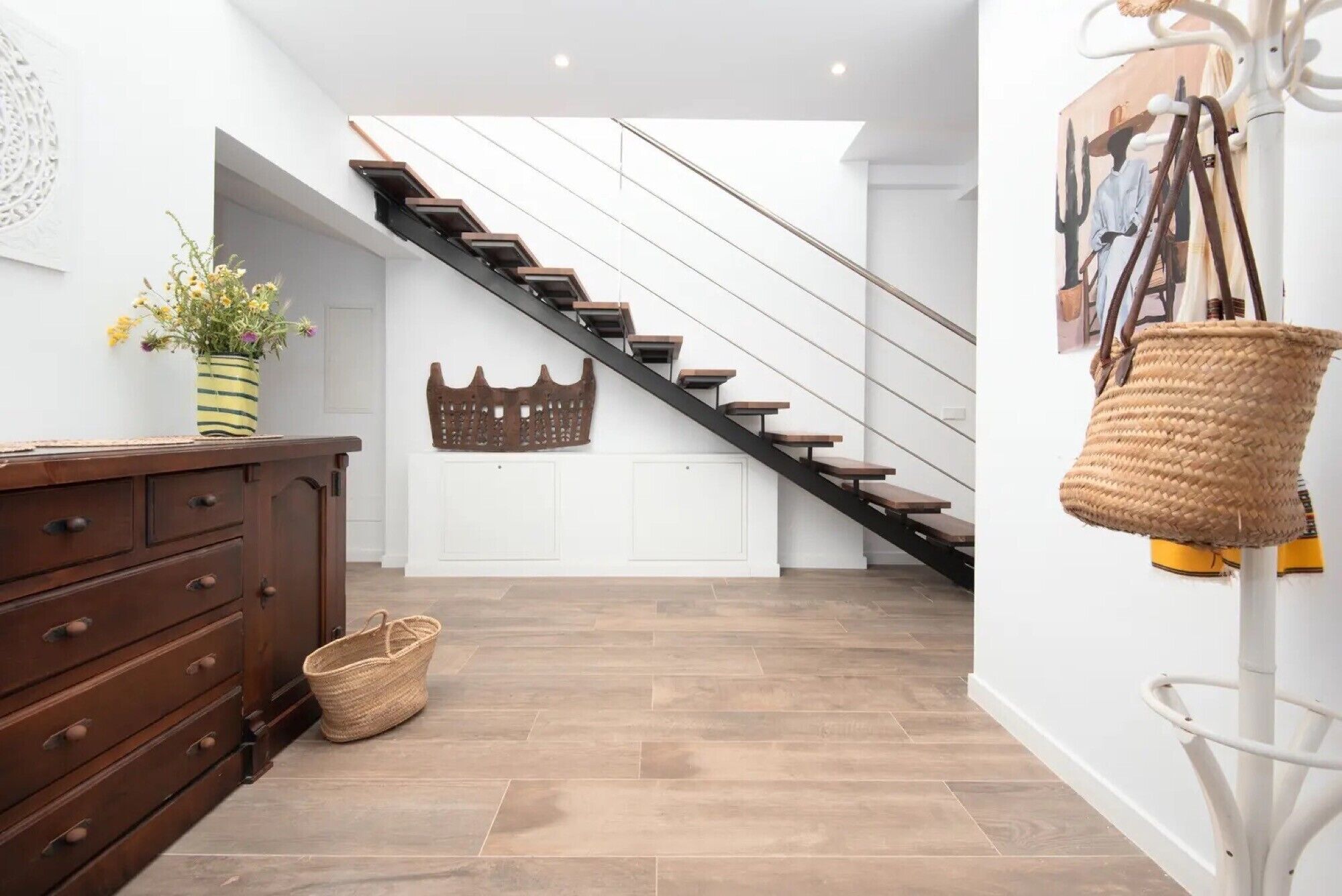 A staircase with a modern design, a wooden dresser, a woven basket, and a wall-mounted artwork.