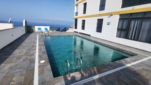 Una piscina con vistas despejadas al océano, un edificio blanco con balcones y una terraza con baldosas de piedra.