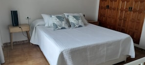 Un lit bien fait, recouvert d’une couette blanche, avec deux oreillers à motifs de feuilles, une table de chevet dotée d’une prise électrique et une armoire en bois.