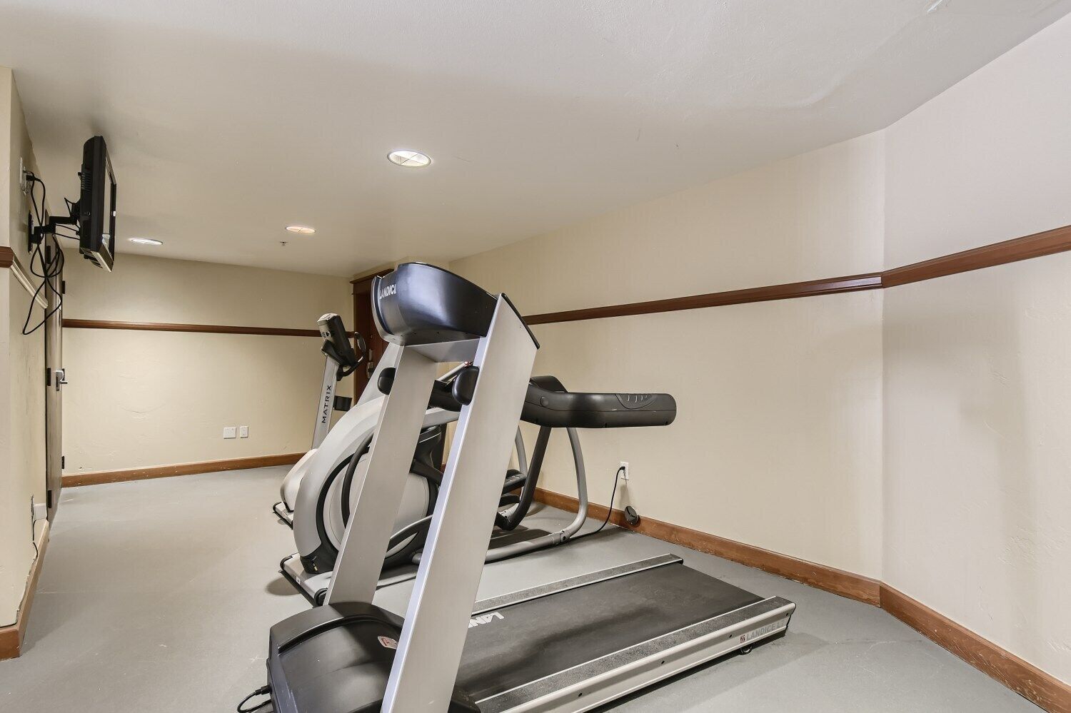 Een sportschool met loopbanden en fitnessapparatuur, een flatscreen televisie aan de muur bevestigd, en een houten vloer met een beige muur.