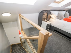 A modern bedroom with a skylight, a bed with pillows, a bedside table with a lamp, and a wooden staircase with glass railings. - The Annex (Ulverston)
