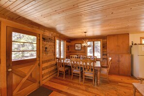Interior de una cabaña de madera con una mesa de comedor, sillas, una araña y un refrigerador.