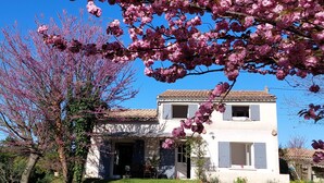 A two-story house with a tiled roof, a front porch, and a garden with blooming pink flowers.