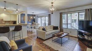 A modern living room with a sofa, coffee table, and TV. There is a dining area with chairs and a table. The room has large windows and a ceiling with a unique design.