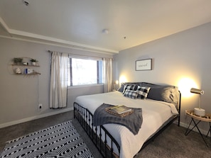A bedroom with a bed, bedside lamp, window with curtains, and a wall-mounted shelf with plants.