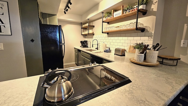 Una cocina moderna con un fogón de inducción negro, un hervidor de acero inoxidable, un tostador y una cafetera. Hay repisas de madera sobre la encimera con frascos y una planta en maceta.