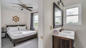 Una camera da letto con un letto, una ventola a soffitto, una decorazione a forma di timone da nave, una finestra e un bagno con un lavandino e uno specchio.