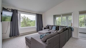 Un soggiorno moderno con un divano modulare grigio, un televisore piatto a parete e ampie finestre con vista sul verde.