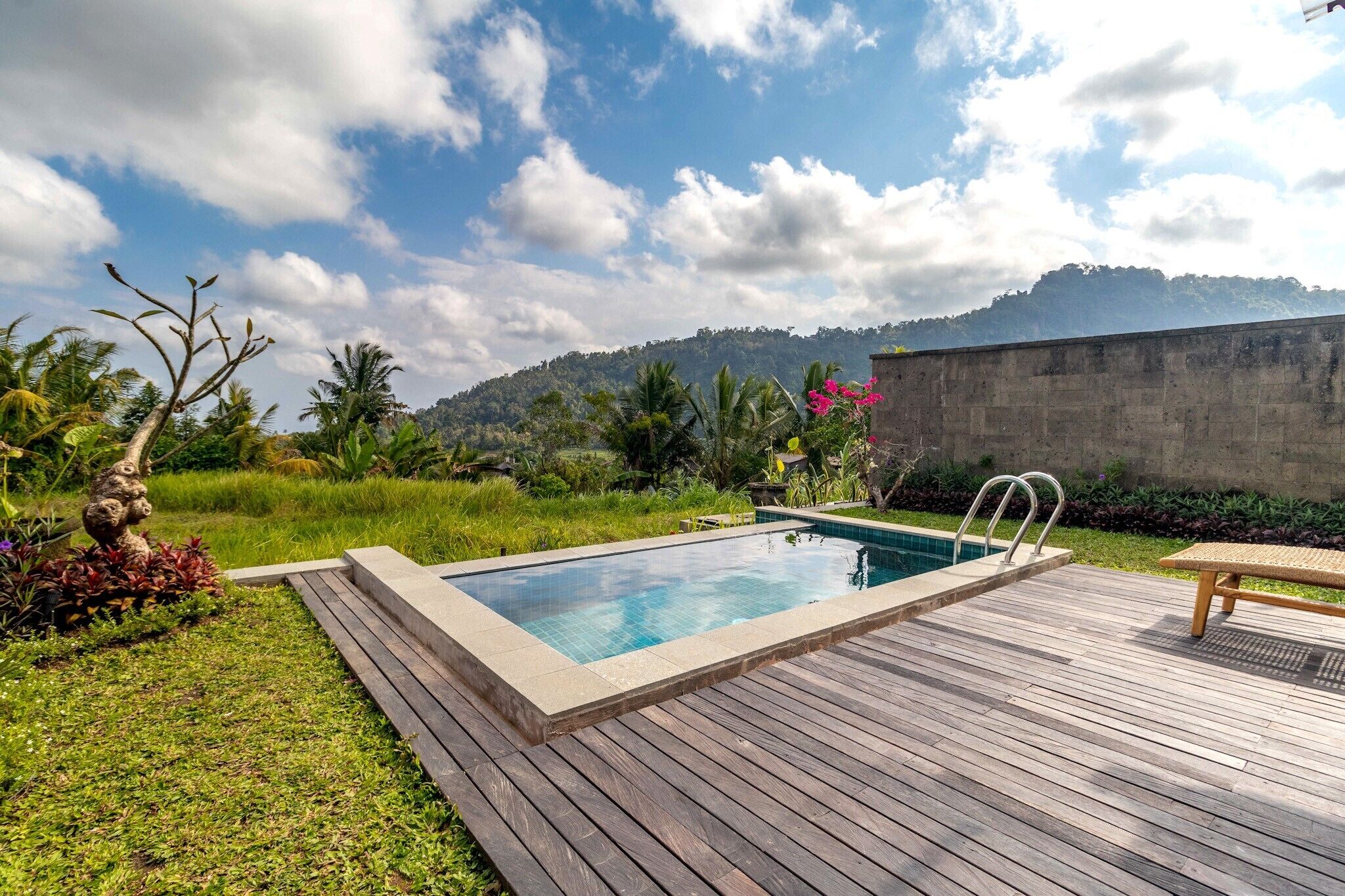 A swimming pool with a wooden deck, surrounded by tropical vegetation and a mountain backdrop.