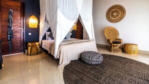 Un dormitorio con cama con dosel, una puerta de madera, una silla tejida y un taburete de madera redondo.