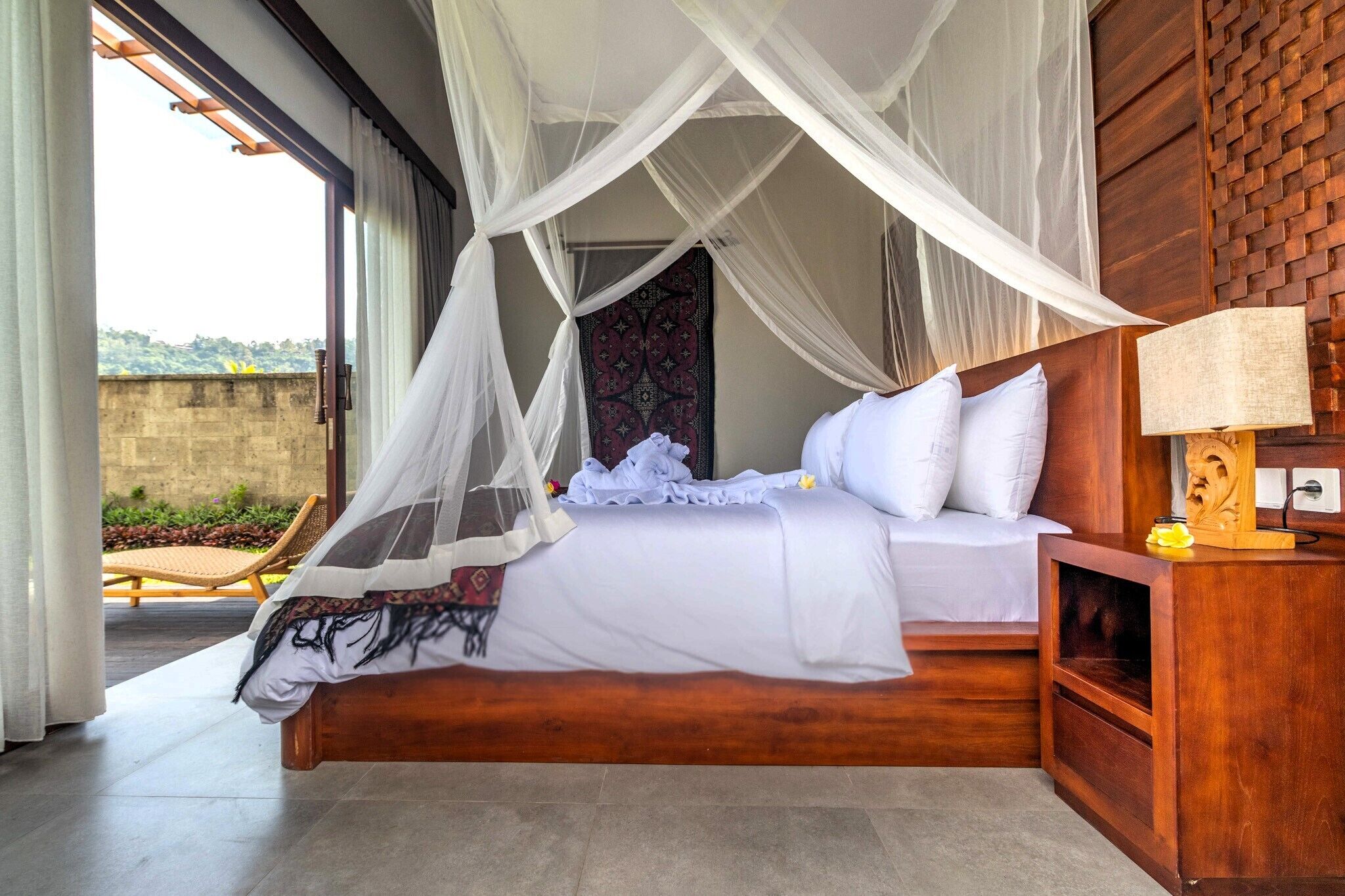 A bedroom with a canopy bed, a wooden headboard, a bedside table with a lamp, and a view of the outdoors.