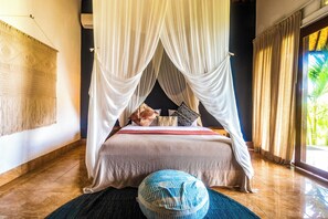 A bedroom with a canopy bed, a woven tapestry on the wall, and a view of greenery outside. - Pelangi Villas Sidemen (Sidemen)