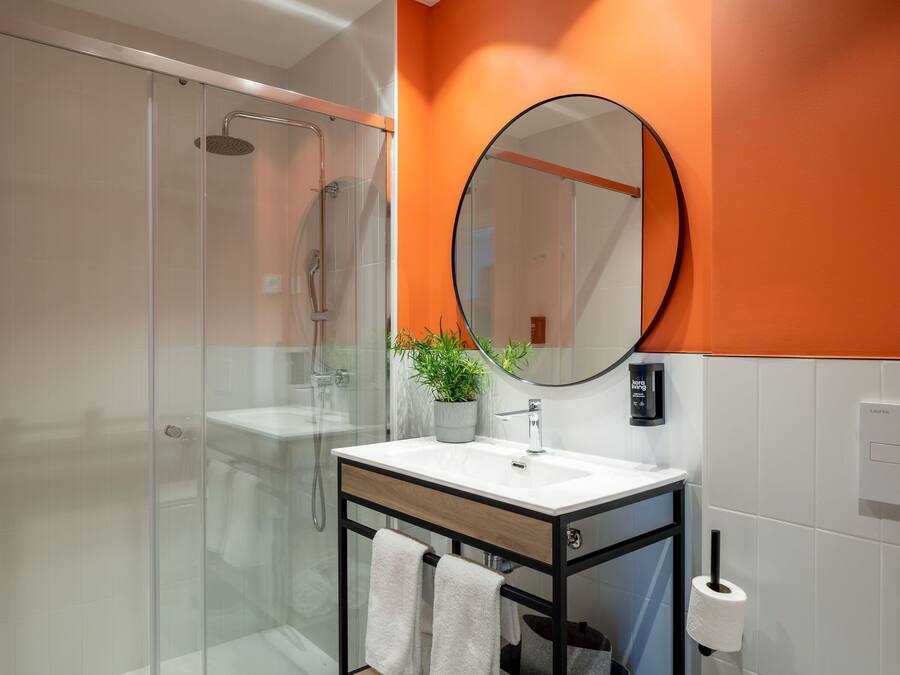 A modern bathroom with a glass-enclosed shower, a white sink with a wooden cabinet, a round mirror, and an orange accent wall.