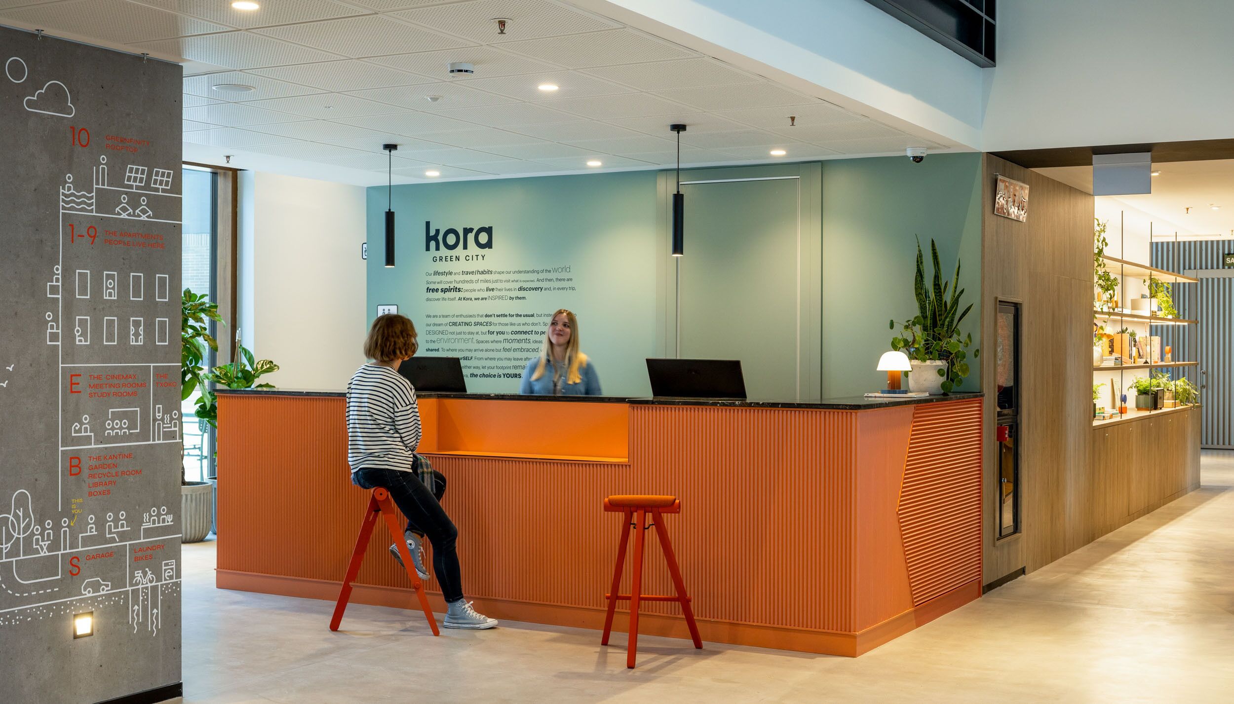 A reception desk with a woman seated behind it, a sign reading "KORA GREEN CITY", and a wall with various icons and text.