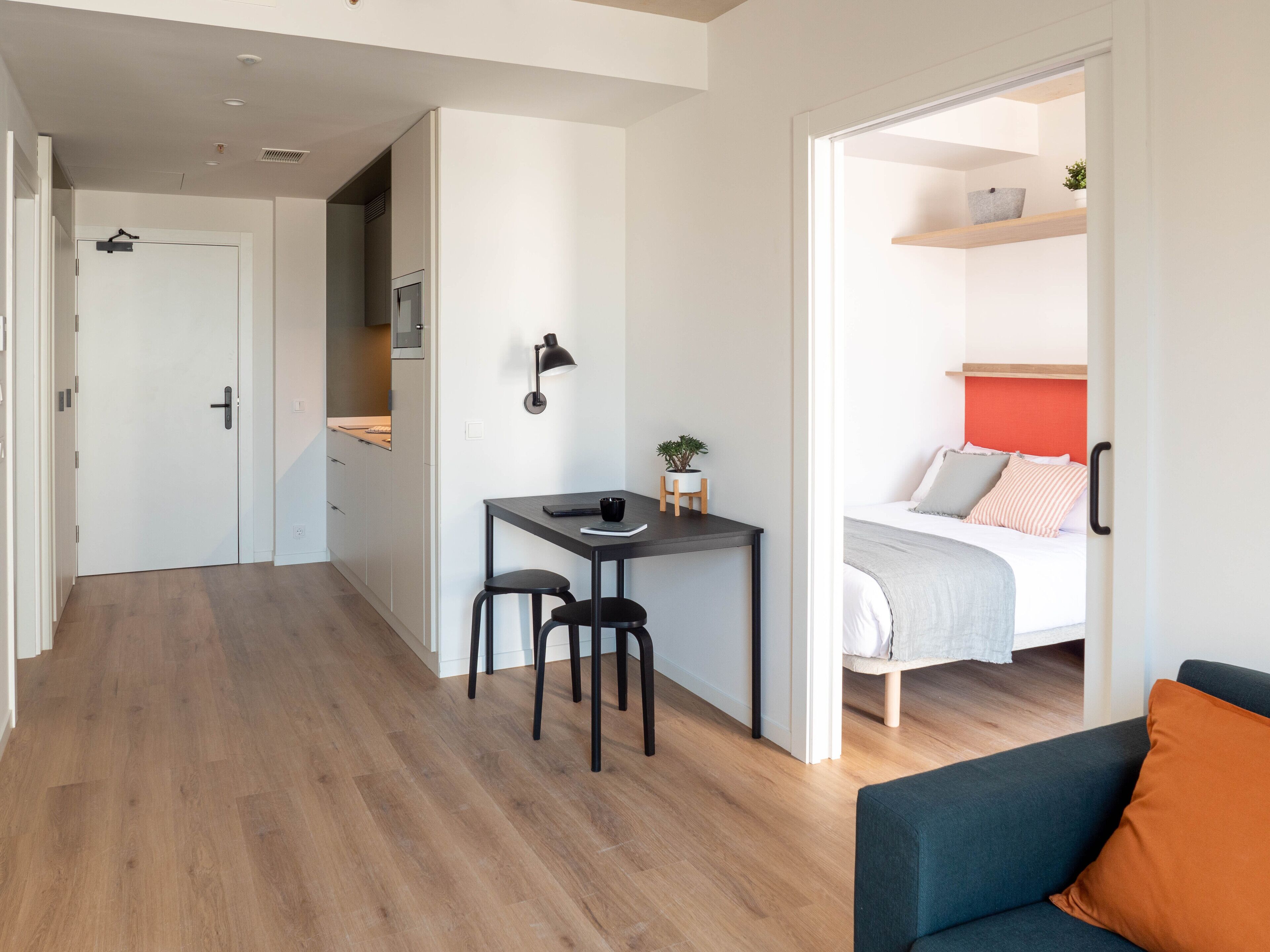 A modern apartment with a kitchen, a dining area with a table and chairs, a bed with a red headboard, and a blue sofa.
