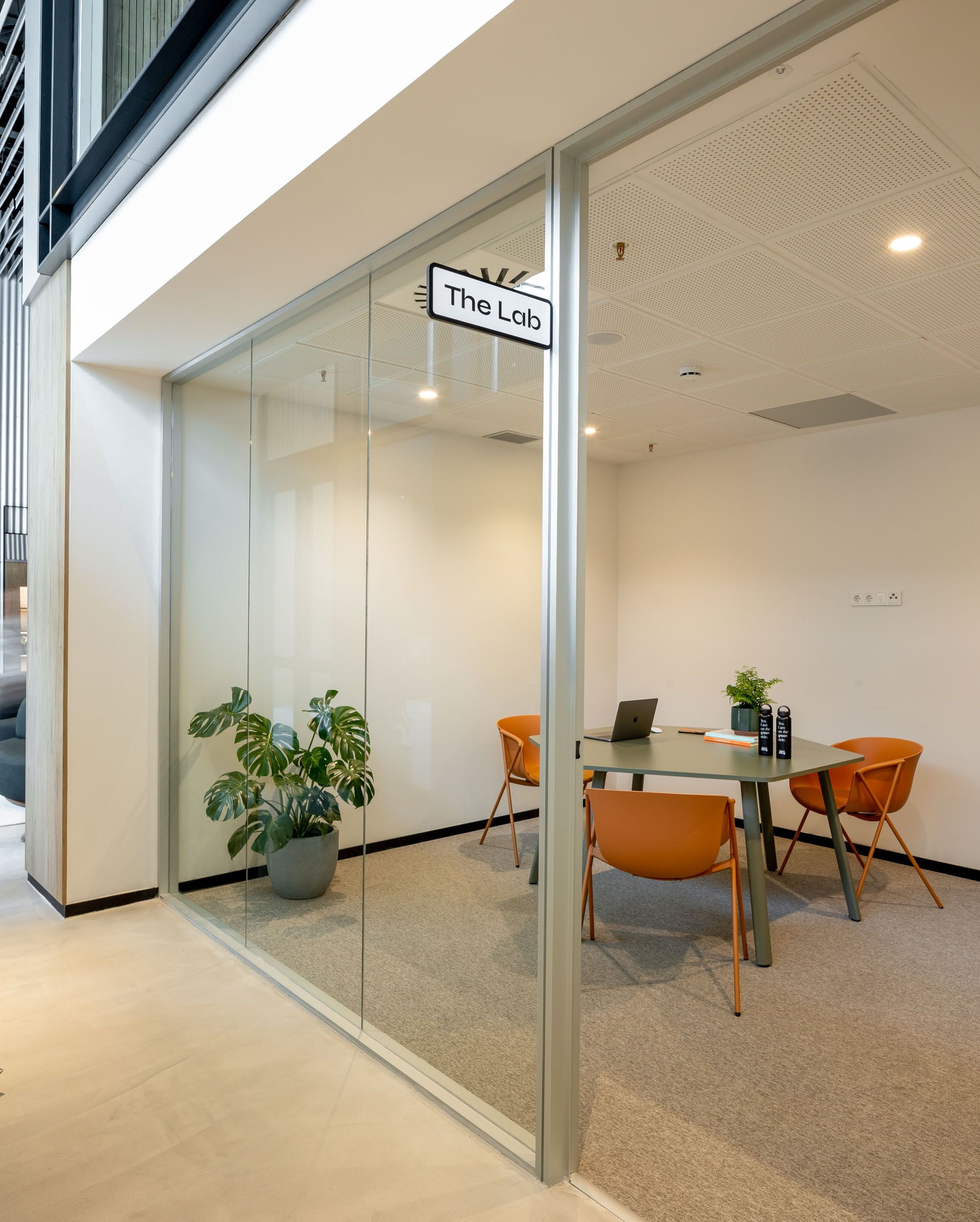 A modern office space with a glass door labeled "The Lab," featuring a meeting table with chairs, a laptop, and a potted plant.