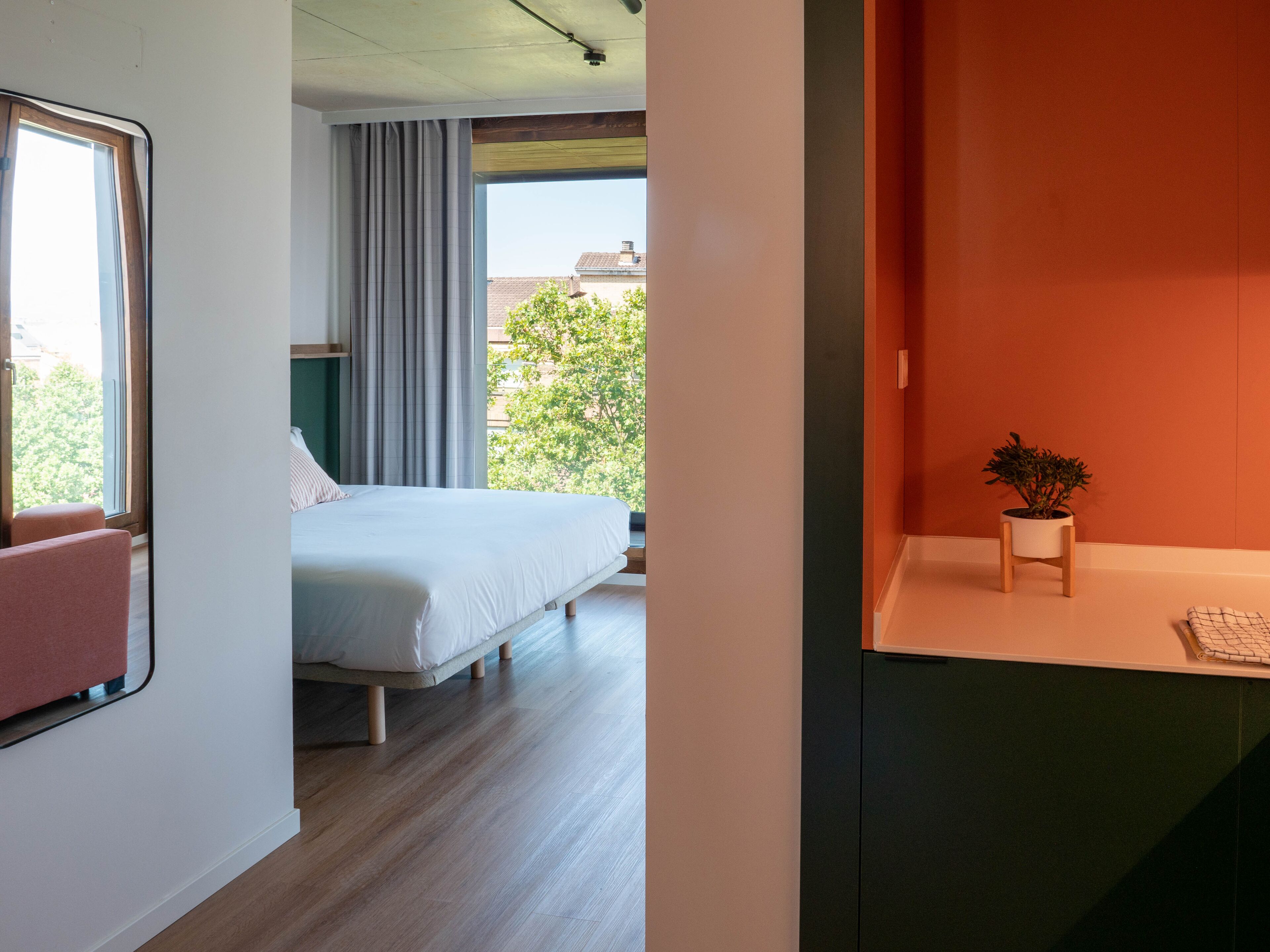 Habitación de hotel moderna con una cama grande, vistas de la ciudad y un pequeño escritorio con una planta.