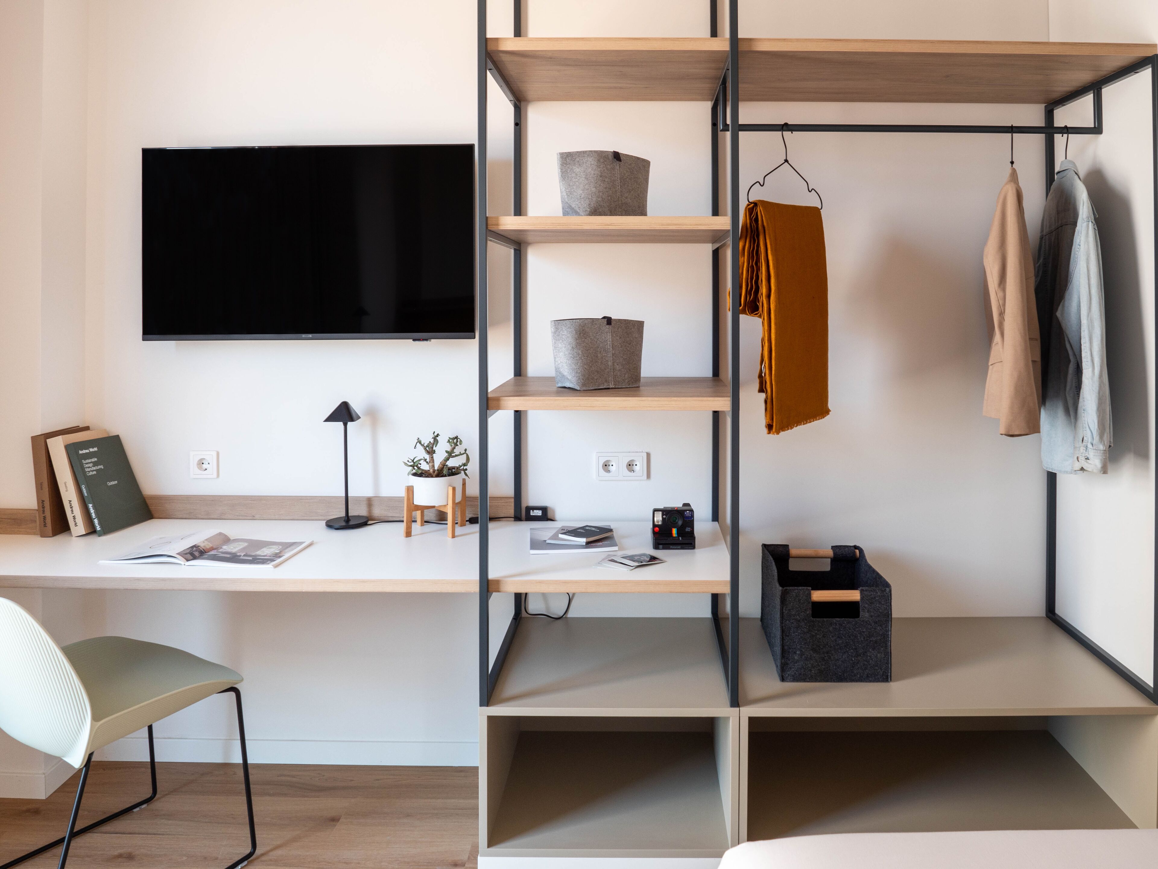 A modern, well-organized room with a flat-screen TV mounted on the wall, a desk with a chair, and a shelving unit with storage boxes and a hanging garment.