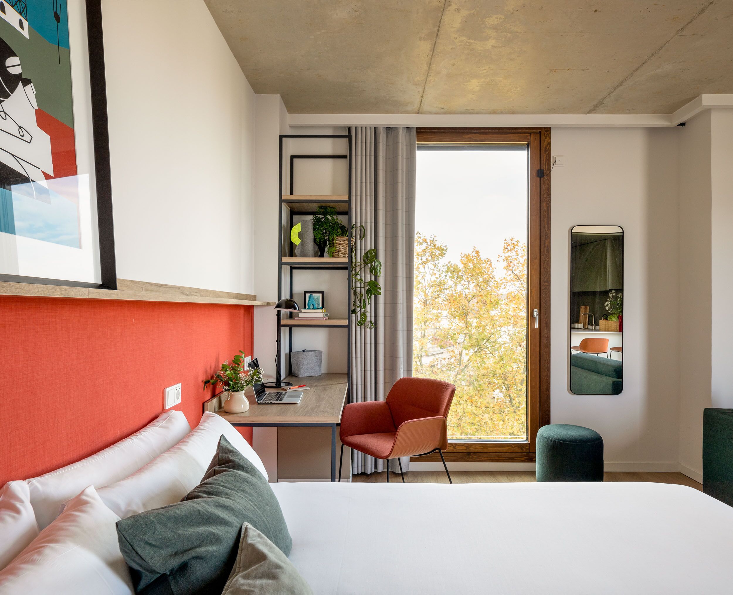 A modern hotel room with a bed, a desk, a chair, a small table, a shelf with plants, and a large window with a view of trees.