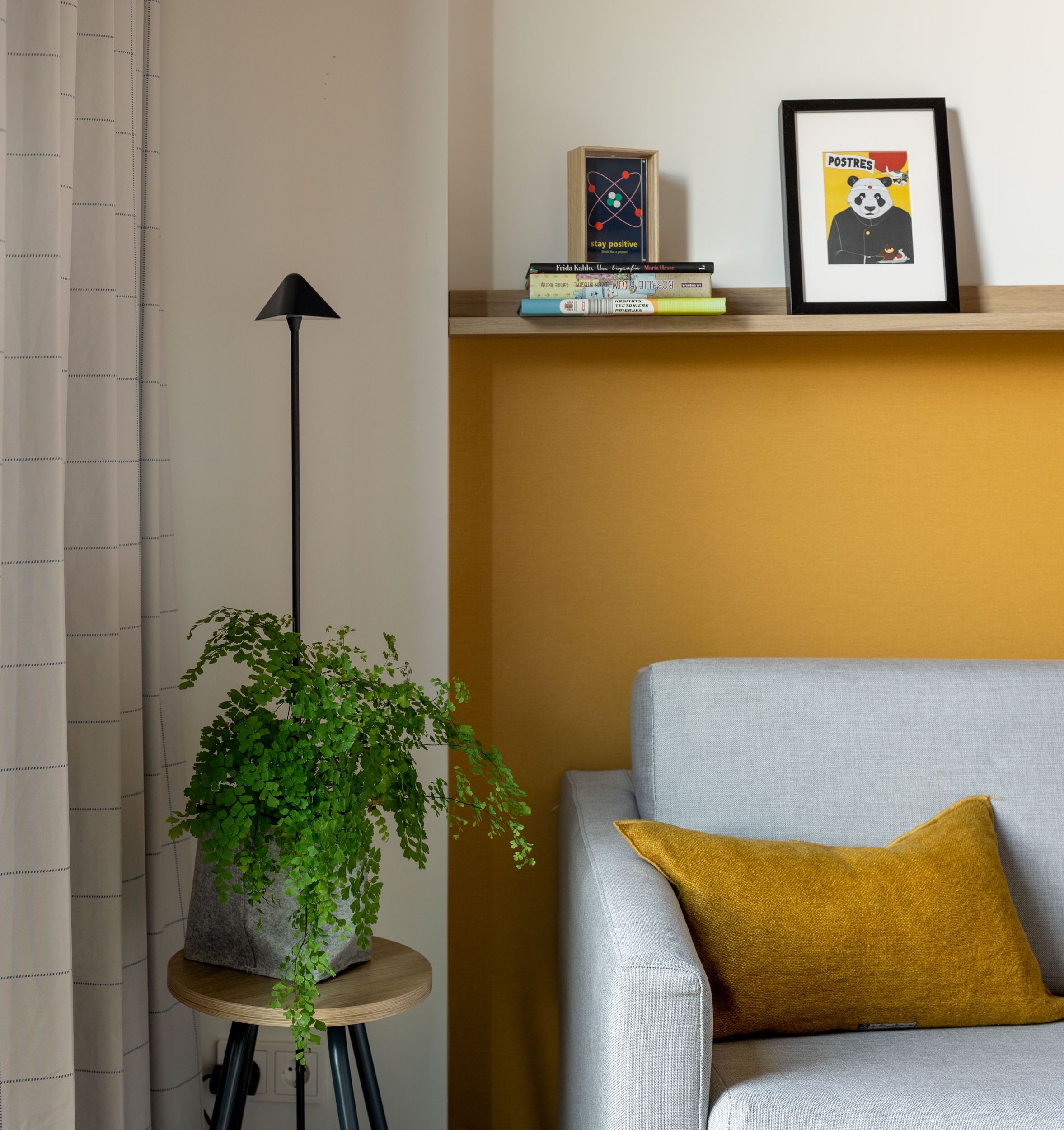A room with a grey sofa, a mustard-colored pillow, a potted plant, a floor lamp, and a shelf with books and a framed poster.