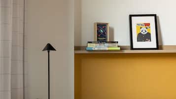 A room with a grey sofa, a mustard-colored pillow, a potted plant, a floor lamp, and a shelf with books and a framed poster.