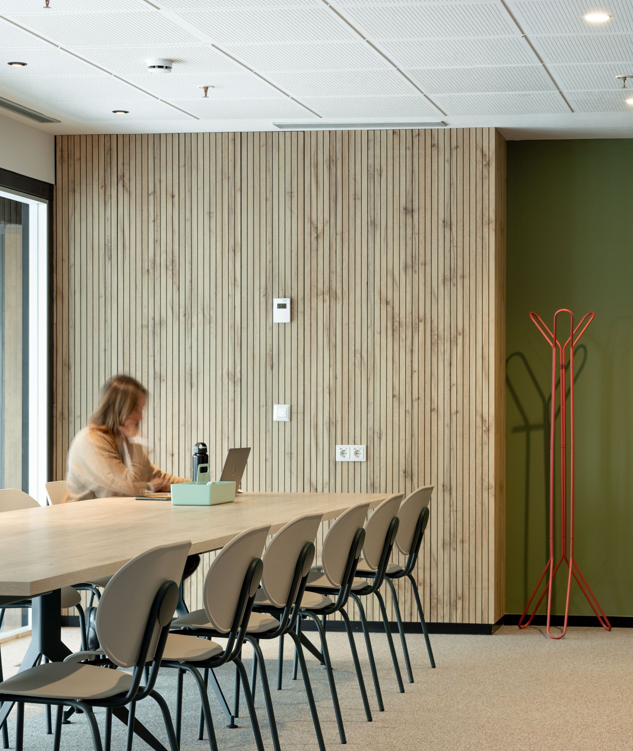 A conference room with a long table, several chairs, a person working on a laptop, a red metal stand, and a wooden panel wall.