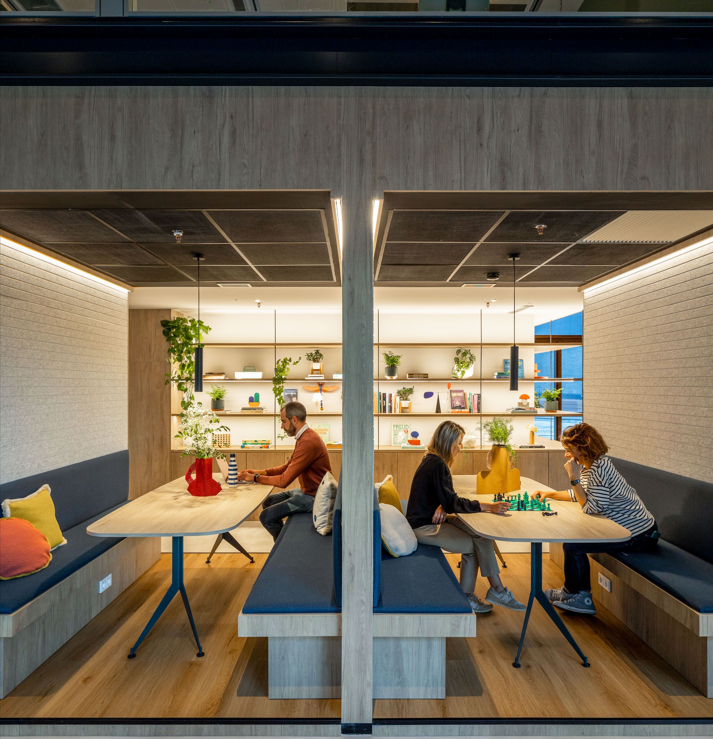 A modern co-working space with a central table, seating areas, and shelves with plants and books.