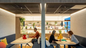 A modern co-working space with a central table, seating areas, and shelves with plants and books.