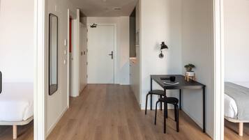 Quarto moderno e bem iluminado, com piso de madeira, uma cama, uma escrivaninha com cadeira e uma mesinha com uma planta.