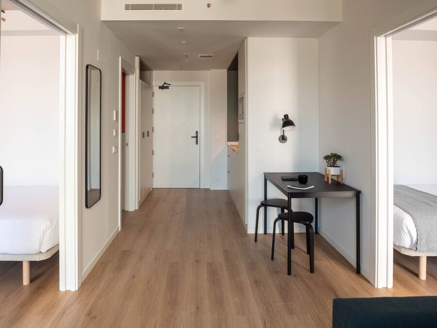Quarto moderno e bem iluminado, com piso de madeira, uma cama, uma escrivaninha com cadeira e uma mesinha com uma planta.