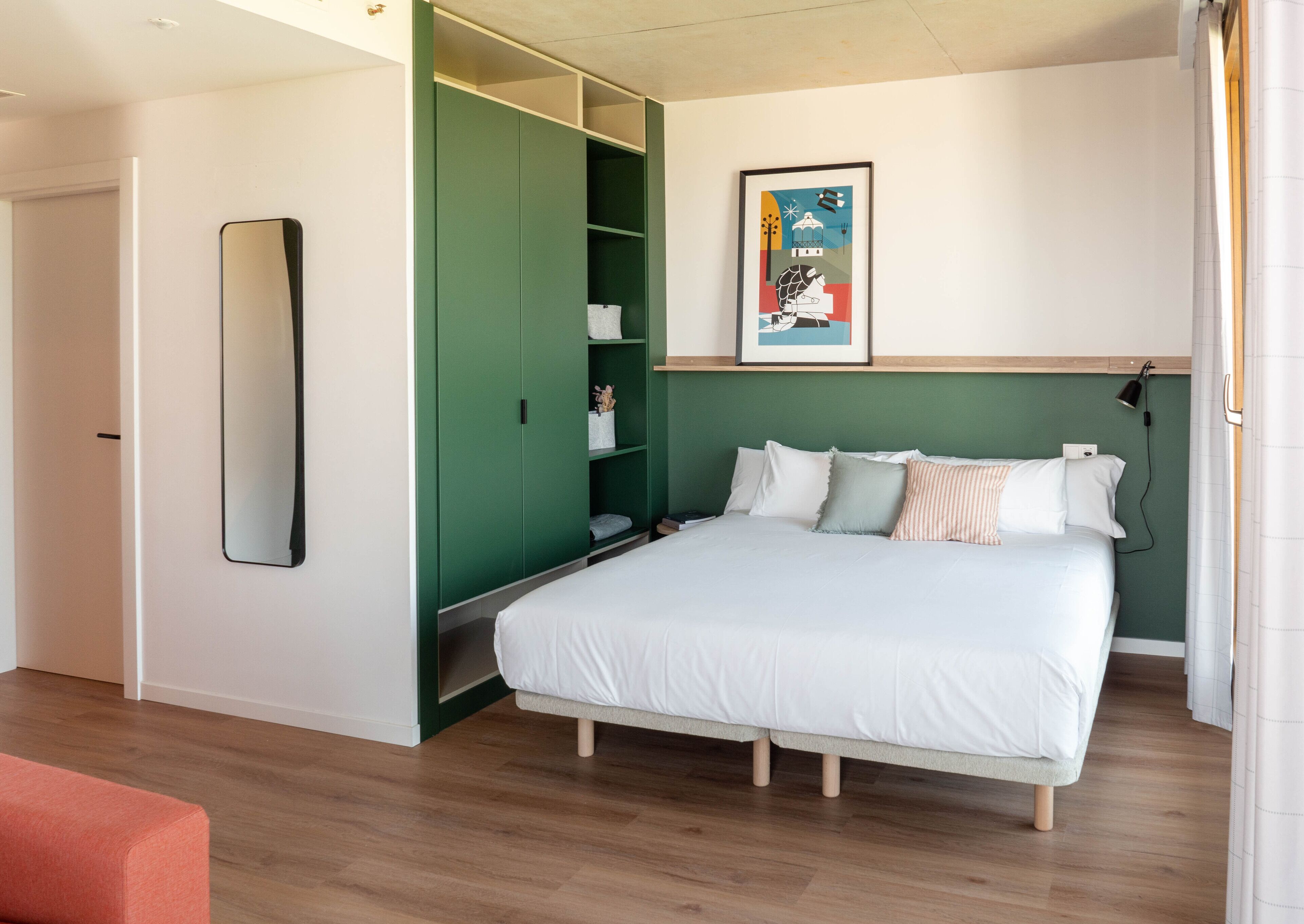 A bedroom with a bed, a green wardrobe, a mirror, and a framed picture on the wall.