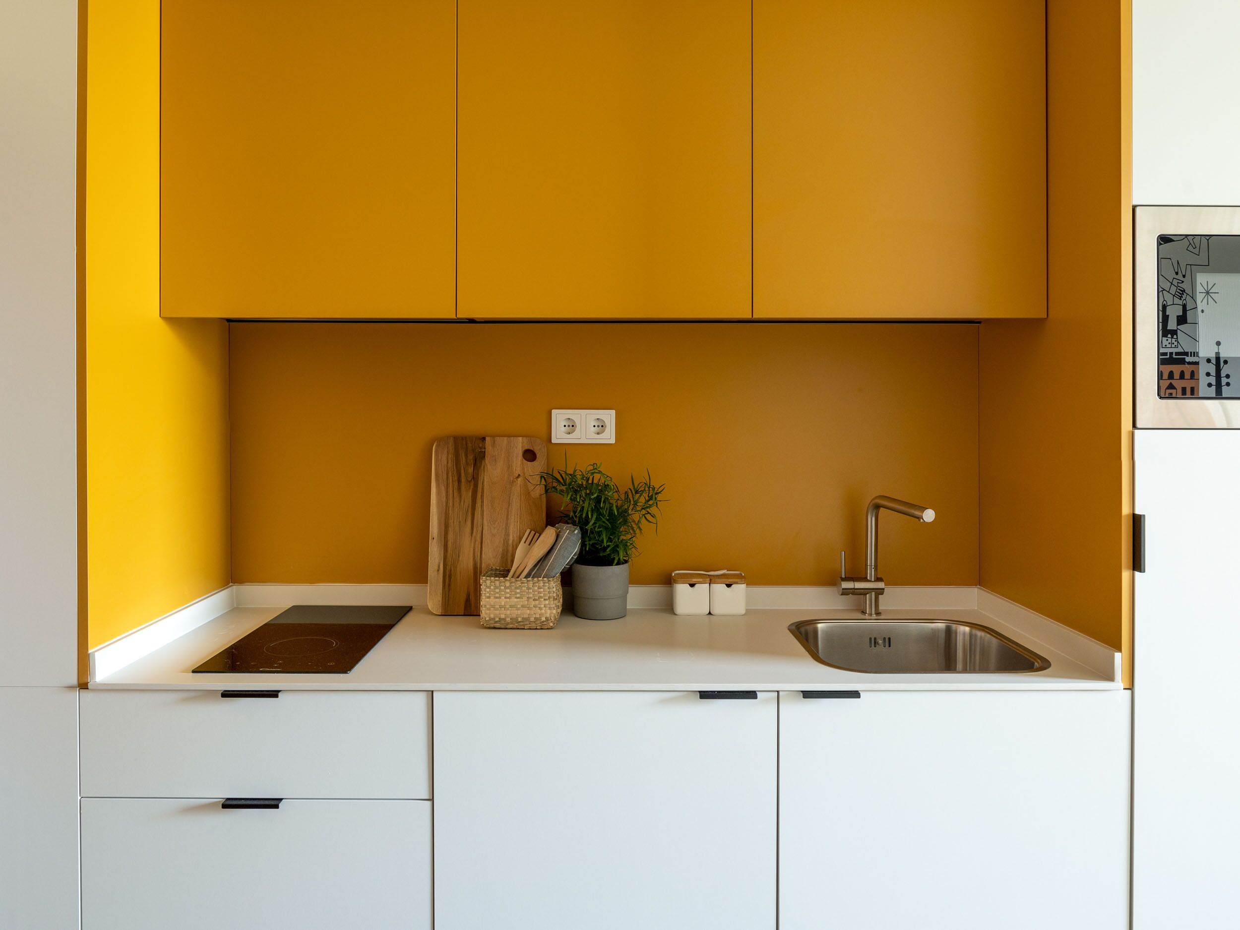 Una cocina moderna con armarios blancos, una pared de acento amarilla, una tabla de cortar de madera, una planta en maceta y un lavamanos de acero inoxidable.