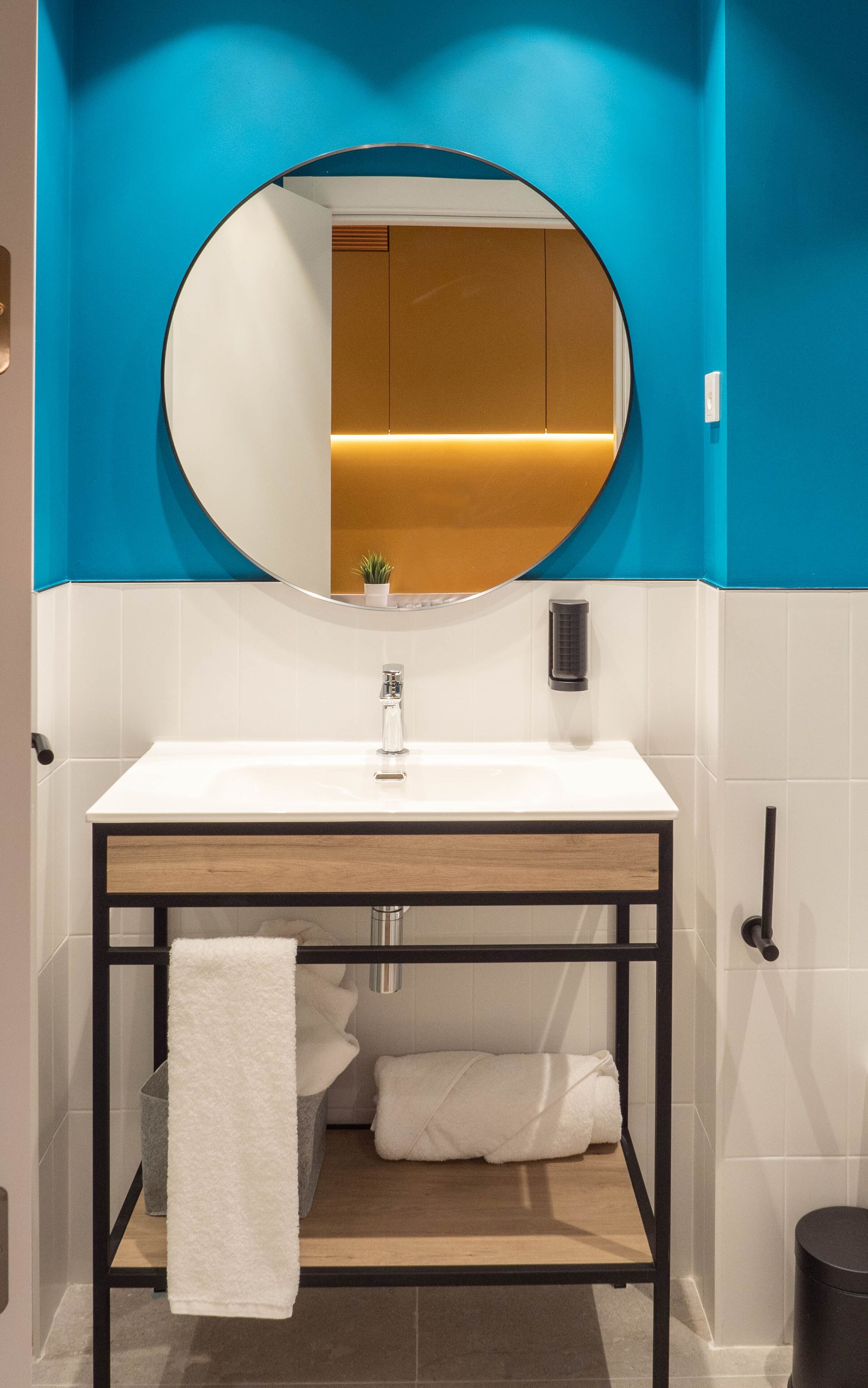Une salle de bain avec un lavabo blanc, un miroir rond et un mur d’accent bleu.