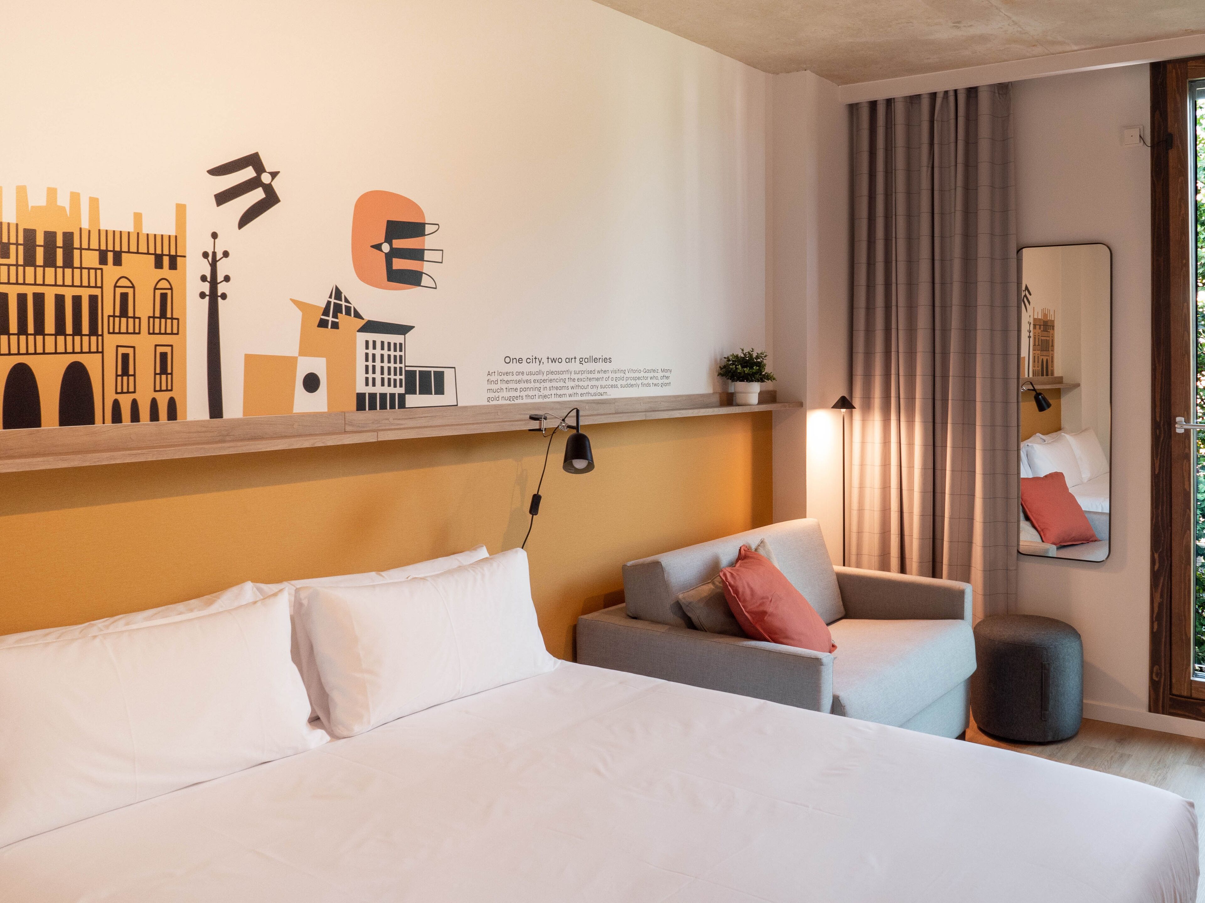 A hotel room with a bed, a sofa, a small table, a mirror, and a wall with art and text.