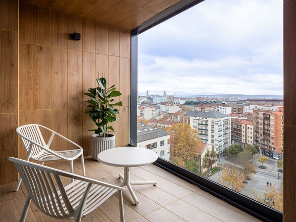 Un balcón con dos sillas blancas y una mesita, con vista a un paisaje urbano con edificios y árboles.