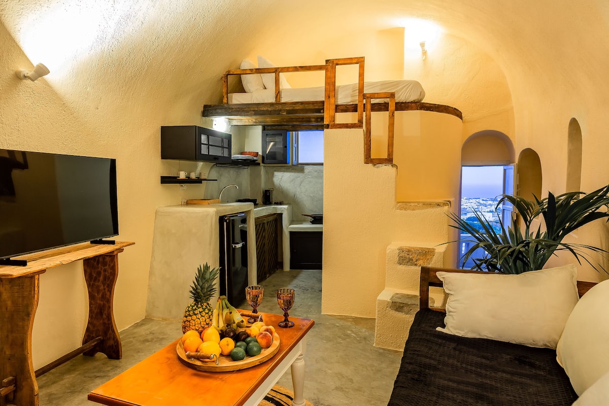 A compact living space with a kitchenette, a television, a dining table with fruit, and a view through an arched window.