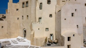 A building with multiple windows and a door, a small outdoor seating area with a table and chairs, and a paved ground with a pattern. - Exi Castle Suites (Santorini)