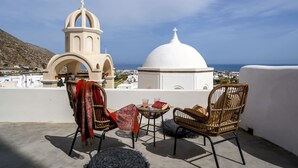 A terrace with wicker chairs, a small table with drinks, and a view of a white church with a dome and bell tower. - Exi Castle Suites (Santorini)
