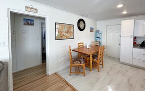 A dining area with a wooden table and chairs, a wall clock, and a kitchen area with cabinets and a refrigerator.