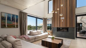 Ein modernes Wohnzimmer mit einer großen Couch, einem Couchtisch und einem Kamin. An der Wand hängt ein Gemälde und große Fenster bieten einen Blick ins Freie.