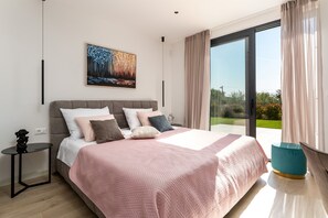 Quarto moderno com uma cama grande, um quadro, uma mesa de cabeceira e vista para o verde através de uma porta de correr de vidro.