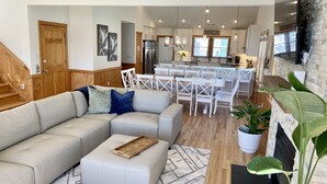 A modern living room with a sectional sofa, a dining area with white chairs, a kitchen with a stone backsplash, and a fireplace.