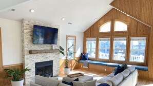 A spacious living room with a stone fireplace, a large flat-screen TV mounted on the wall, a built-in bench with cushions, and large windows offering a view of the outside.