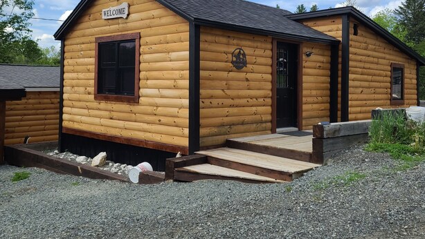 Una cabaña de madera con un letrero que dice 'Welcome', un porche pequeño y una entrada de grava.
